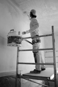 A person steadily painting a living room wall with a roller while furniture is covered and painter’s tape lines the trim.