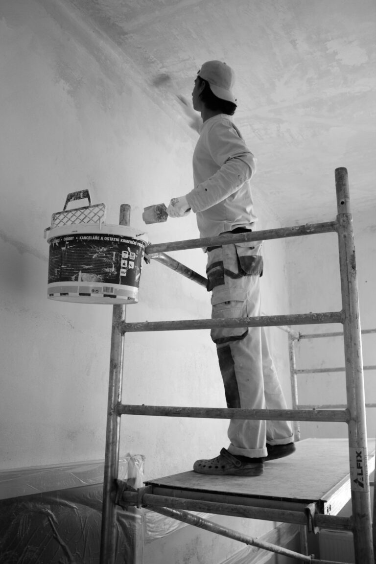 A person steadily painting a living room wall with a roller while furniture is covered and painter’s tape lines the trim.