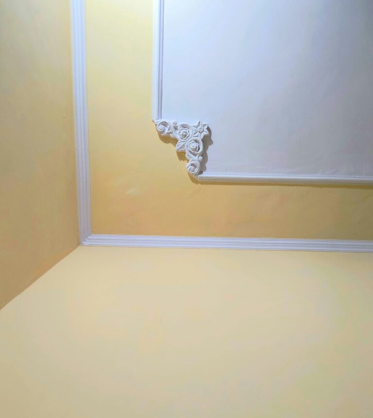 Painter using an angled brush to apply white paint along detailed wood molding beside a wall.